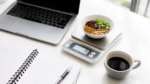 A closed laptop, kitchen scale with weighed ingredients, open notebook, pen and cup of black coffee on a bright desk