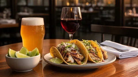 A glass of craft beer and a glass of red wine beside a plate of grilled fish tacos on a bistro table
